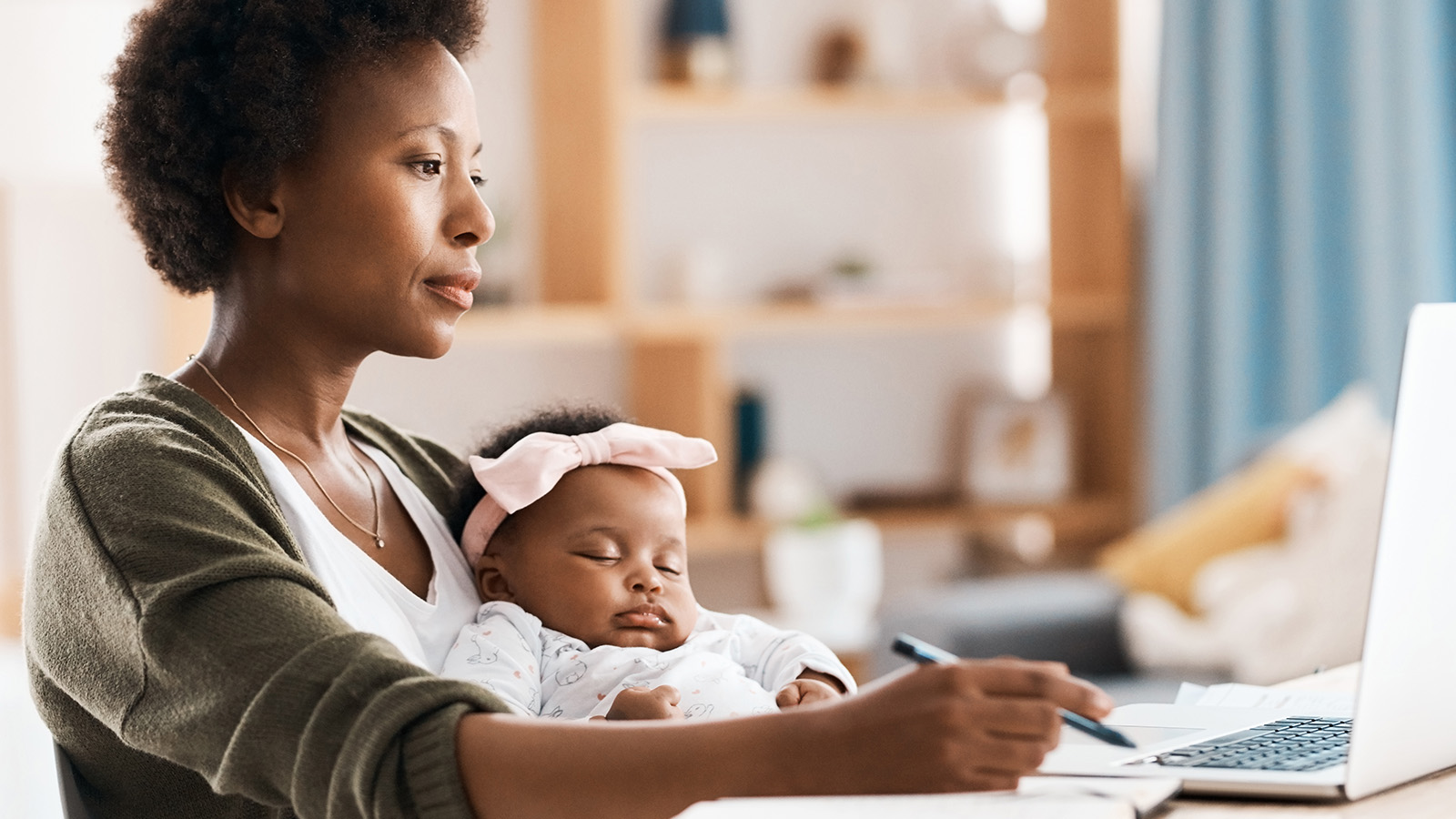 A mother holds her baby while writing notes at a desk, surrounded by a bright and organized living space.