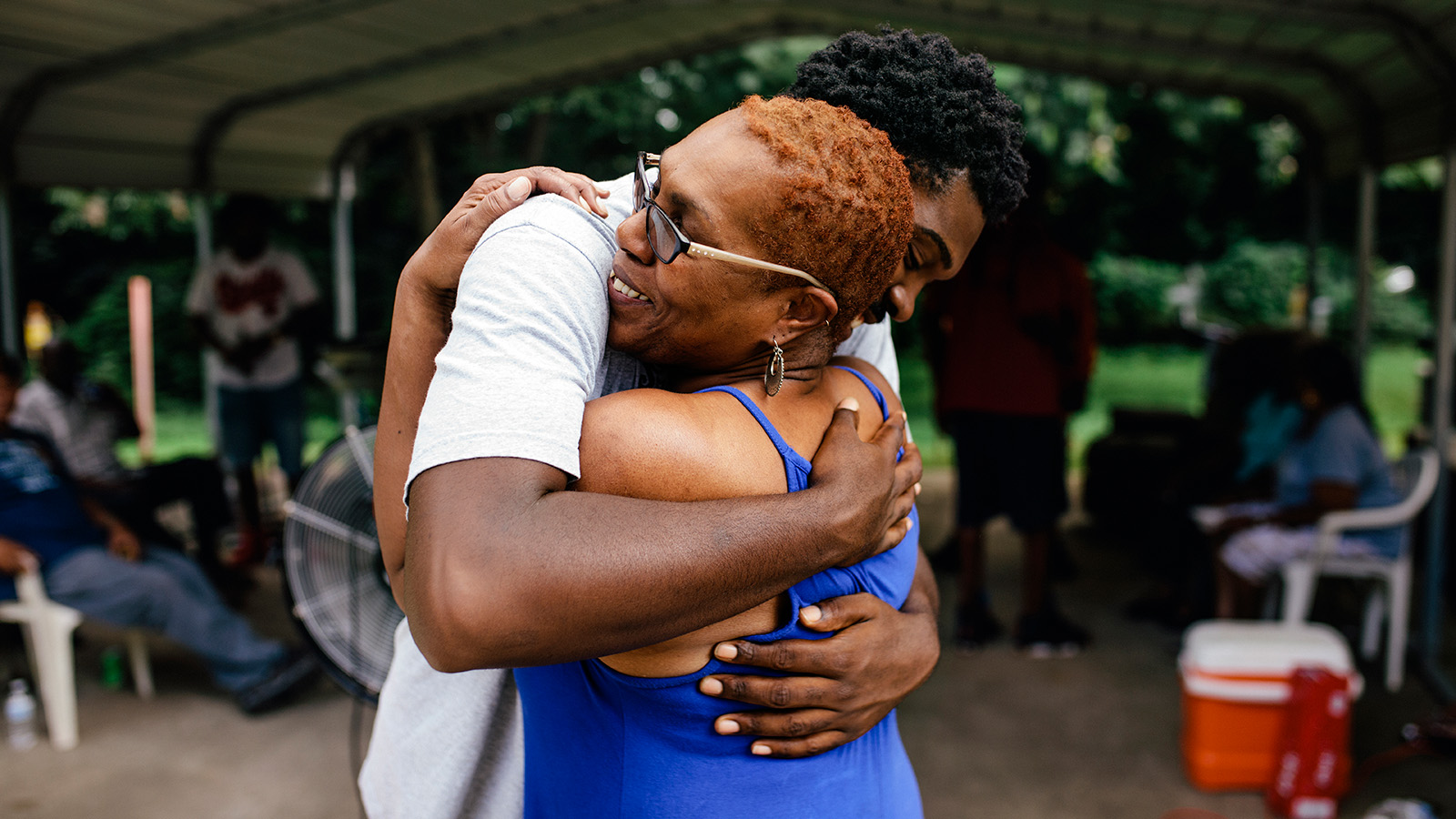 Two people embrace warmly outdoors, surrounded by friends and family, conveying a sense of love and support.