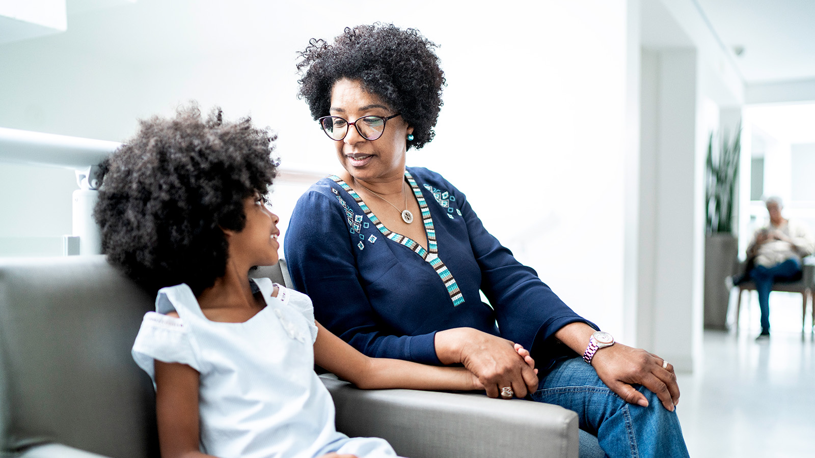 An adult and child sit together in a waiting area, holding hands and looking at each other with affection.