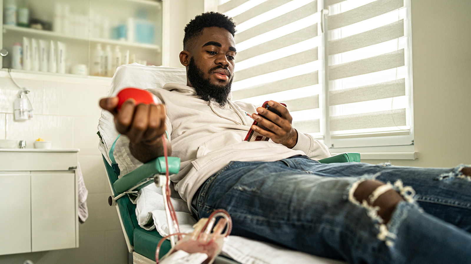A person relaxes in a medical chair, donating blood while holding a phone, with medical supplies in the background.