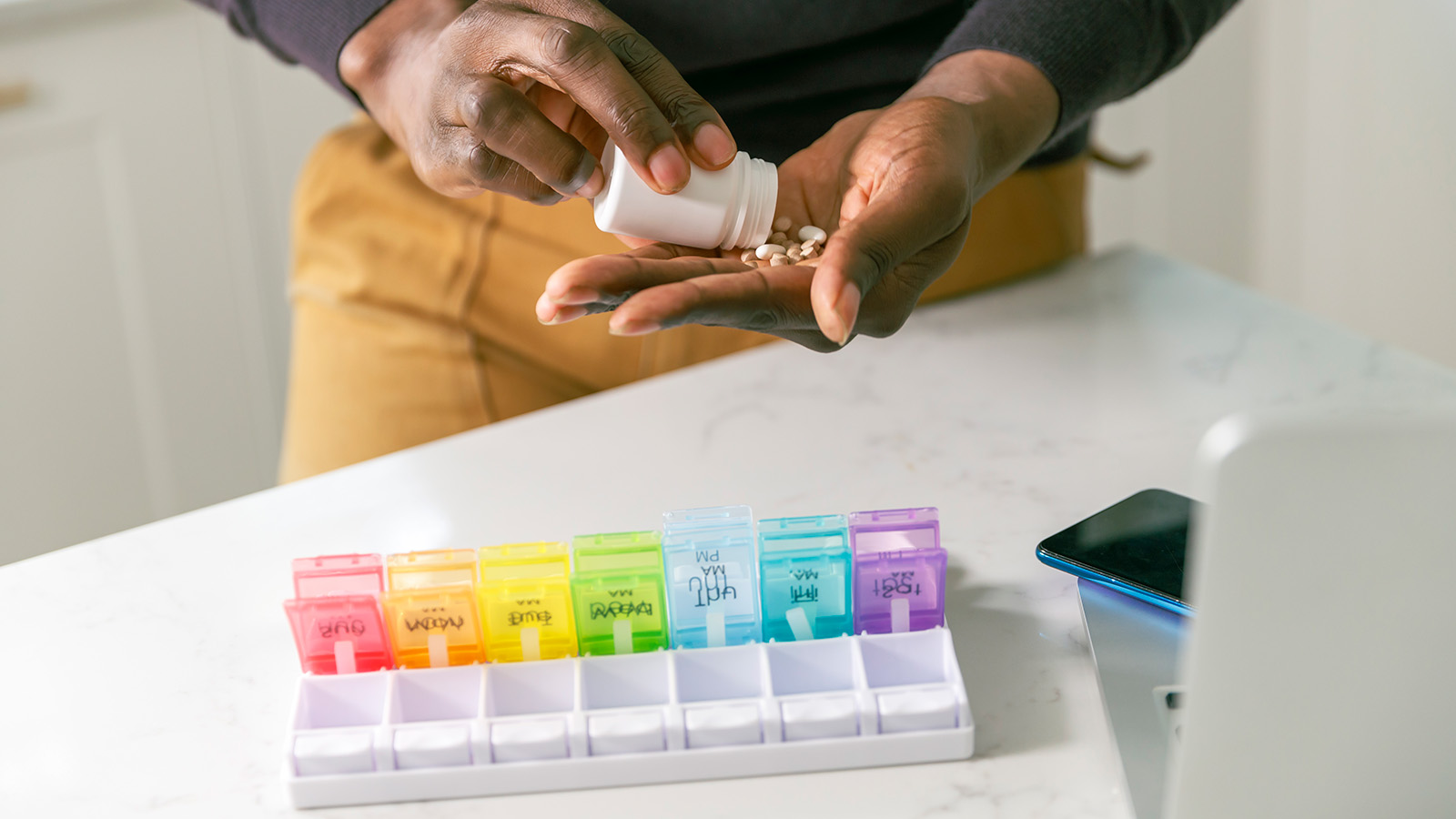 A person pours pills from a bottle into their hand, with a colorful weekly pill organizer nearby on a countertop.
