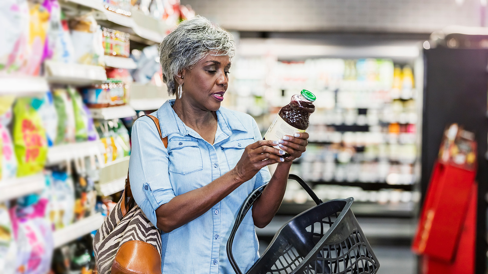 A woman in a grocery store examines a bottle while holding a shopping basket.