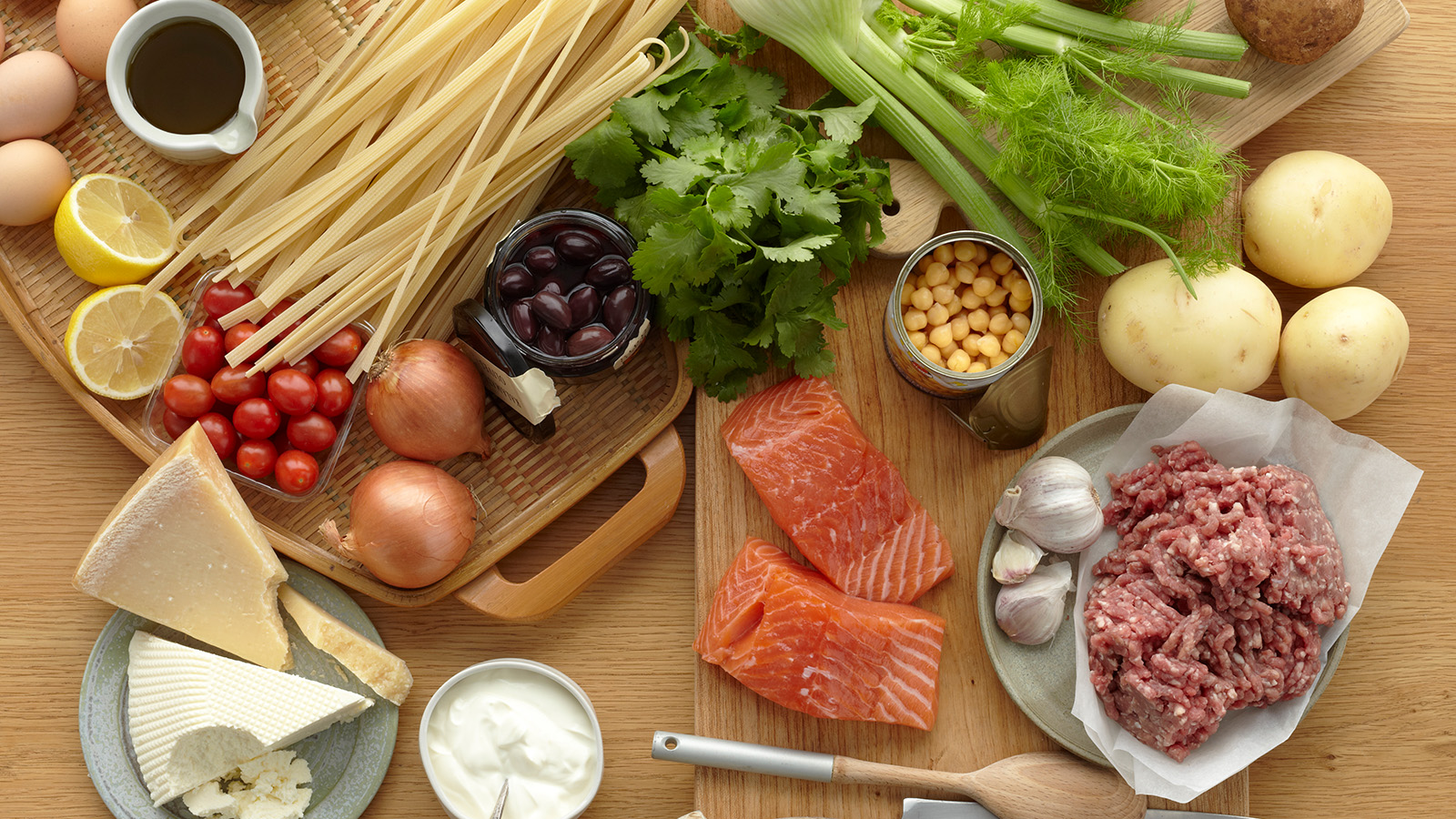 A variety of fresh ingredients, including vegetables, pasta, and fish, are arranged on a wooden surface.