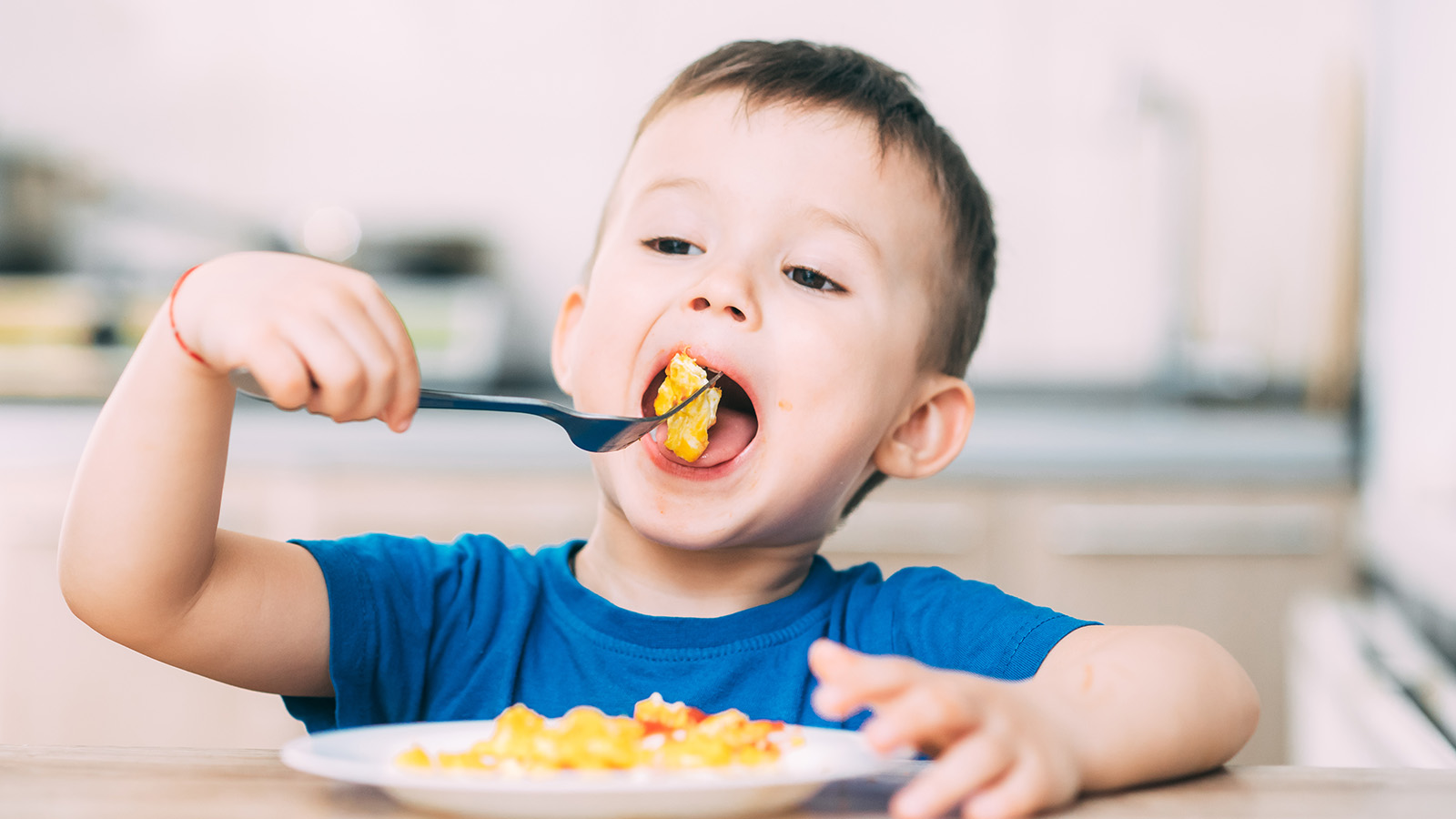 A child in a blue shirt eats pasta with a fork, sitting at a table in a kitchen setting.