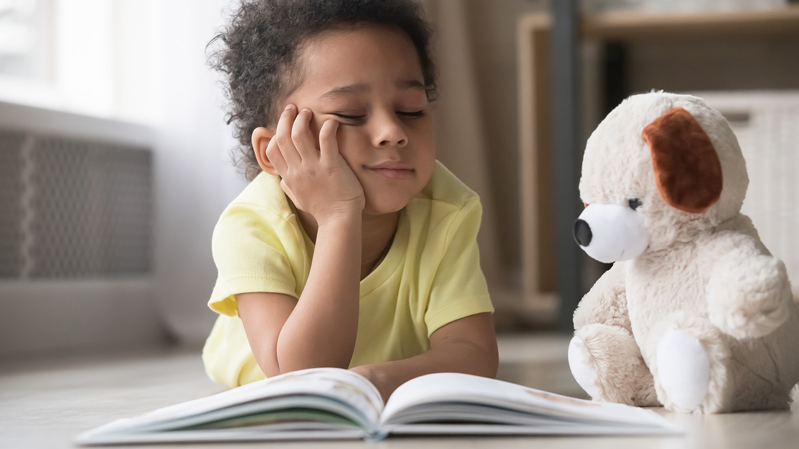 A young child lies on the floor, resting their chin on their hand, looking at a book with a stuffed dog nearby.