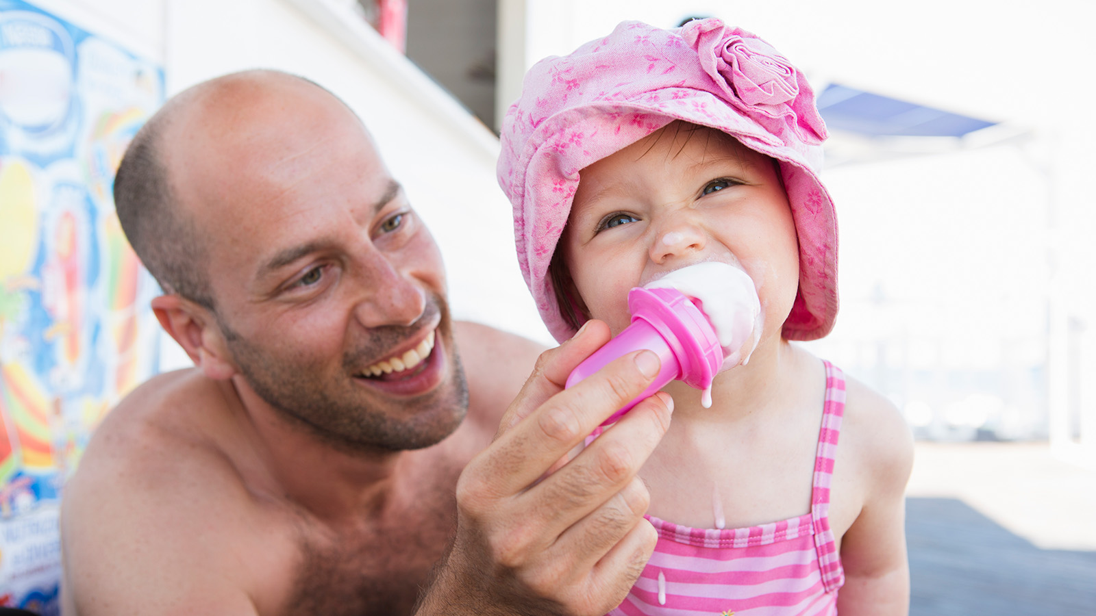 A man and a young girl share an ice cream treat outdoors, with the girl wearing a pink hat and striped swimsuit.