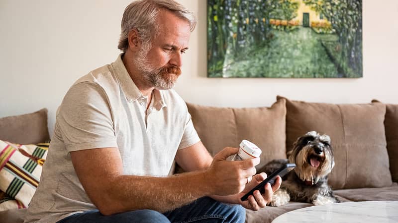 A person on their sofa beside their dog, checking their prescription on their cell phone.