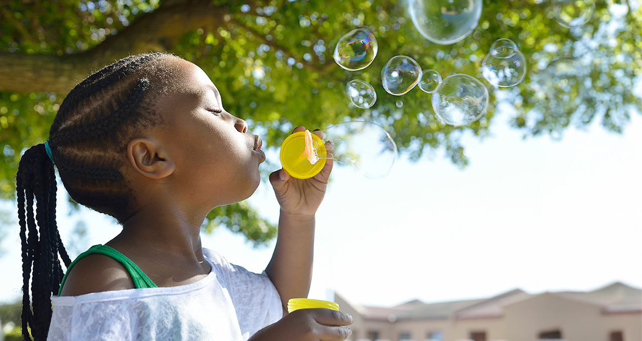 A girl blows bubbles outdoors, with sunlight filtering through trees, enjoying a playful moment in the park.