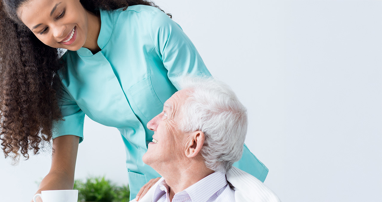 A caregiver serves a drink to an elderly man, both engaged in conversation in a bright, modern setting.