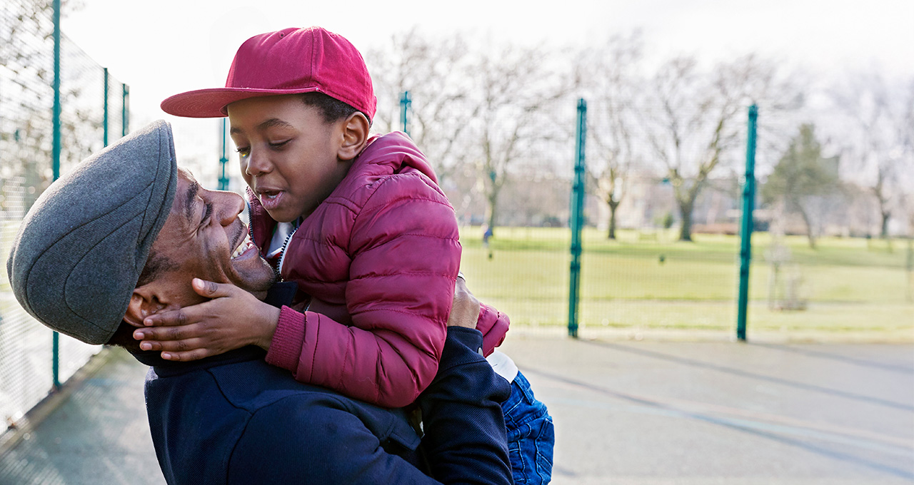 A father joyfully lifts his child in the air, both smiling, in a park setting with trees in the background.