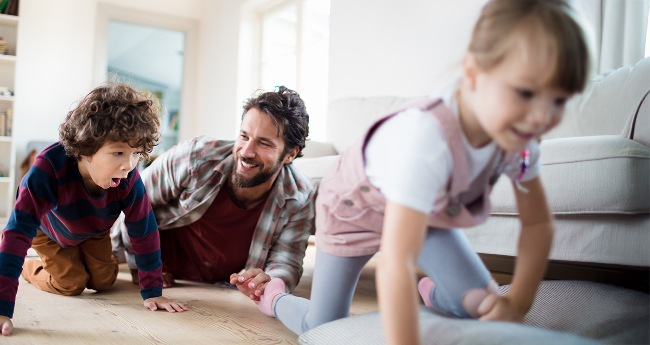 A father plays on the floor with two children, engaging in a fun activity in a bright living room.