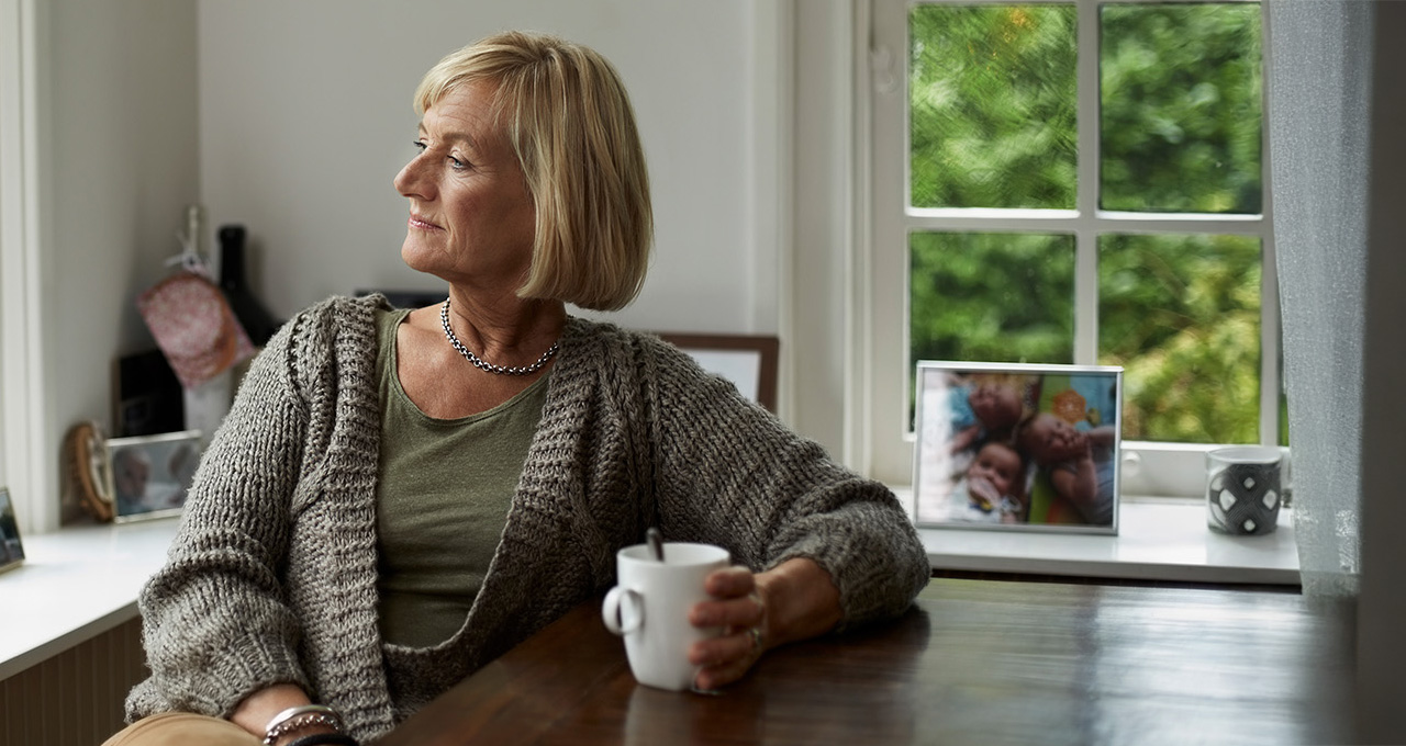 A woman sits at a table with a cup, looking out a window with greenery and family photos nearby.