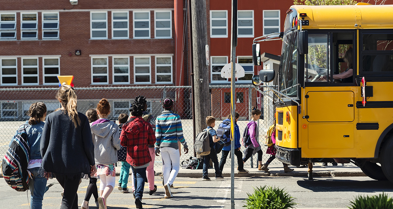 A group of children walks towards a yellow school bus in front of a brick school building.