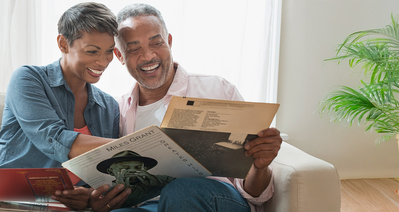 A couple sits together on a couch, sharing a vinyl record, surrounded by natural light and a plant.