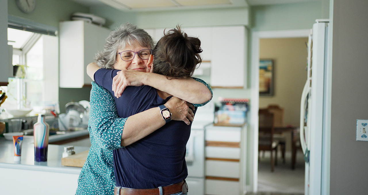 Two people embrace in a cozy kitchen, surrounded by homey decor and natural light streaming through the window.