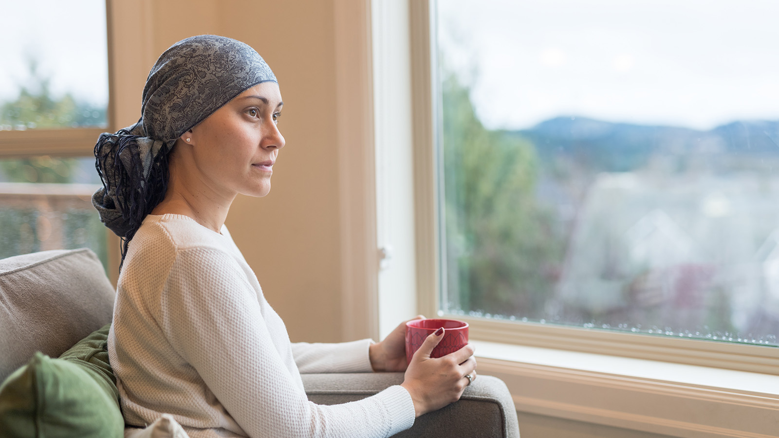 A woman sits on a couch, holding a red mug and gazing out a window at a scenic view.