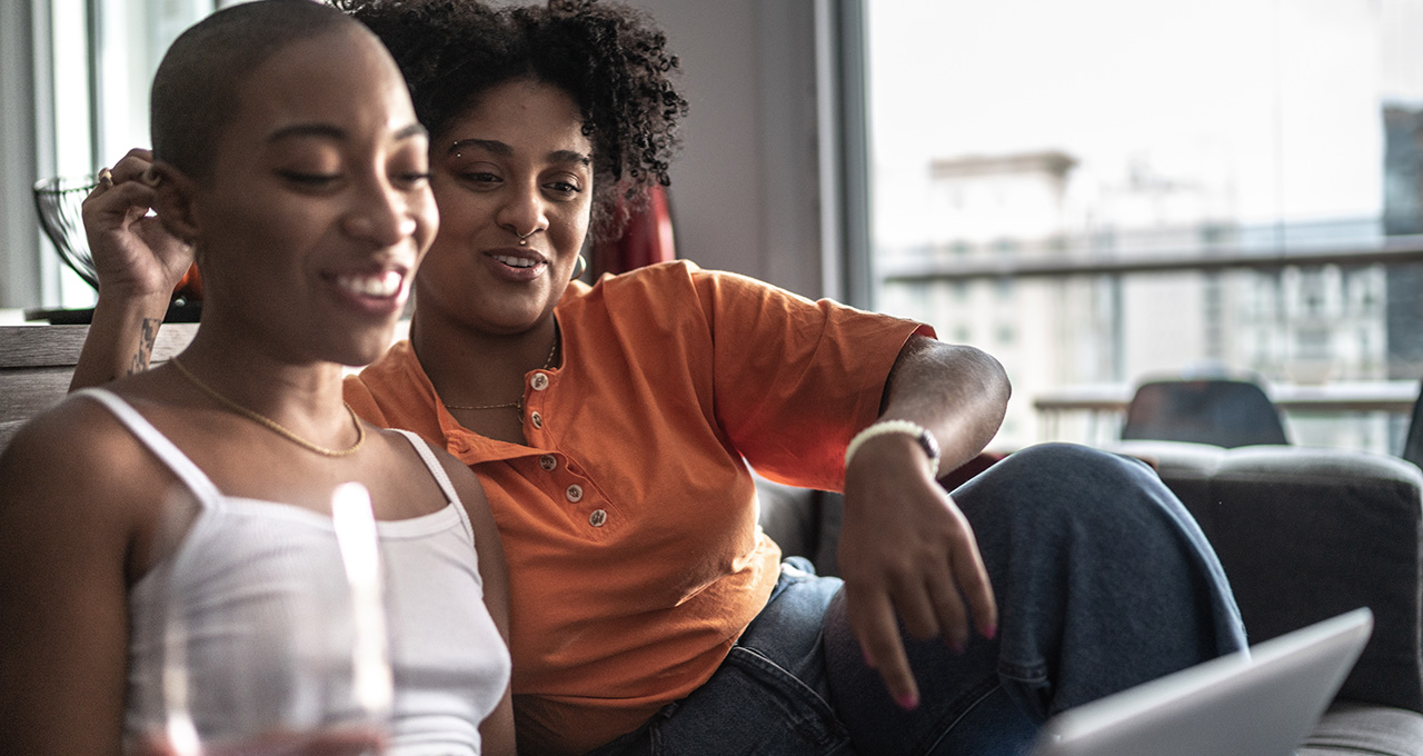 Two friends relax on a couch, one with a drink, sharing a moment of connection and laughter indoors.