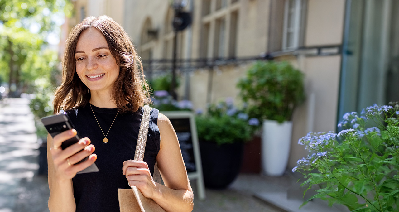 A woman stands outdoors, holding a smartphone and looking at it, surrounded by greenery and bright sunlight.