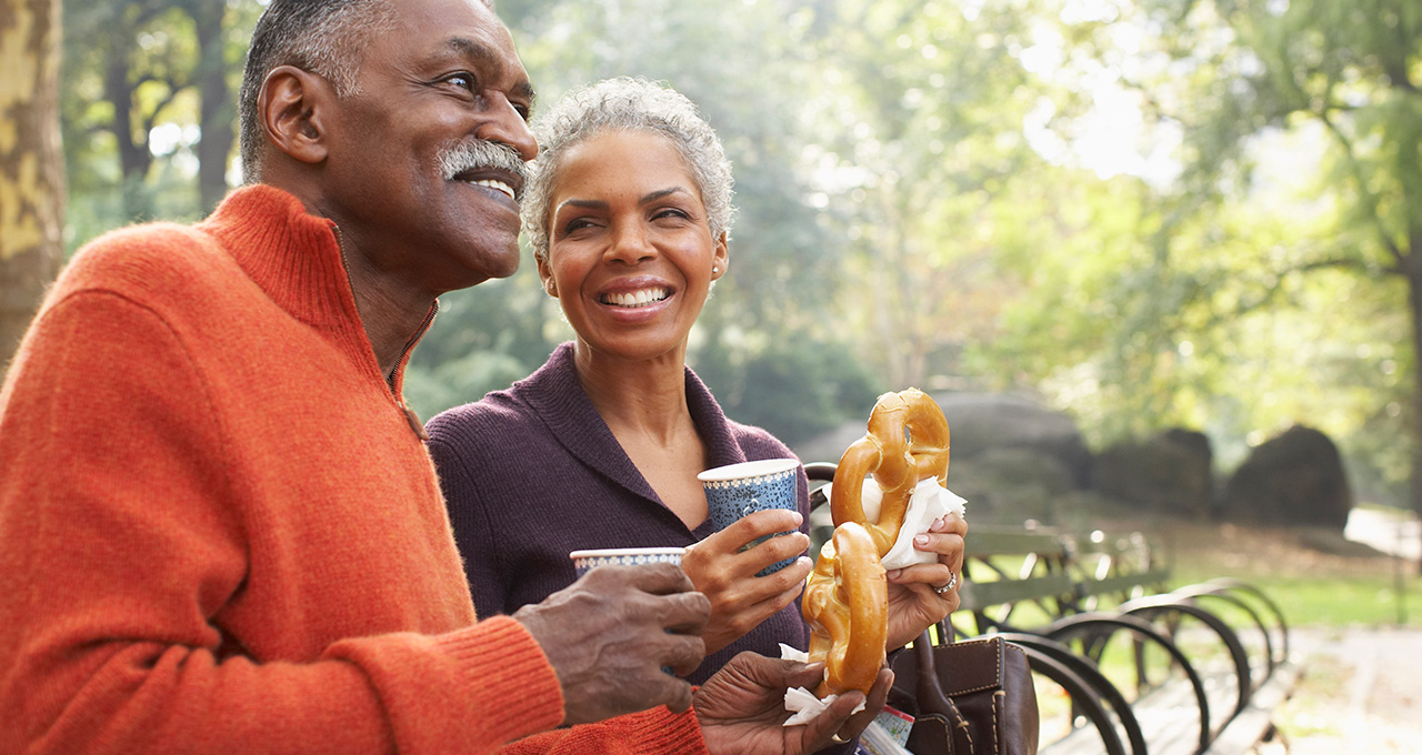 A couple enjoys snacks and drinks while standing in a park, surrounded by trees and a peaceful outdoor setting.