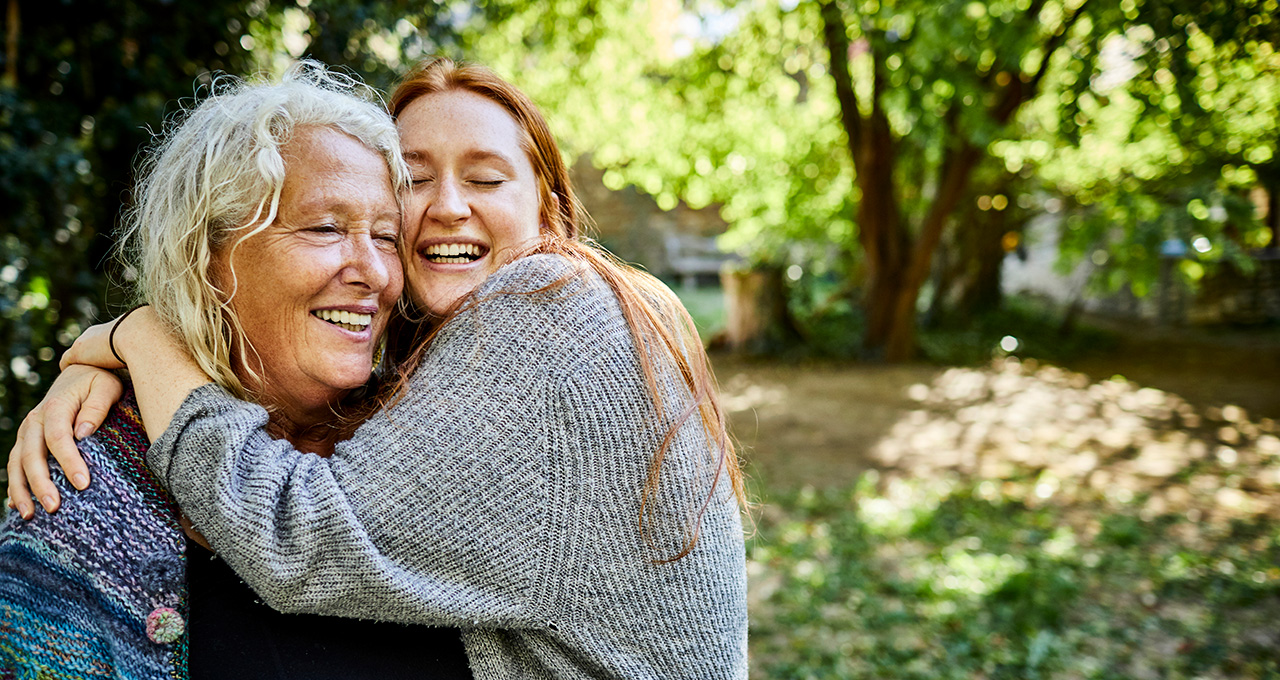 Two women embrace warmly outdoors, surrounded by lush greenery and soft sunlight filtering through the trees.