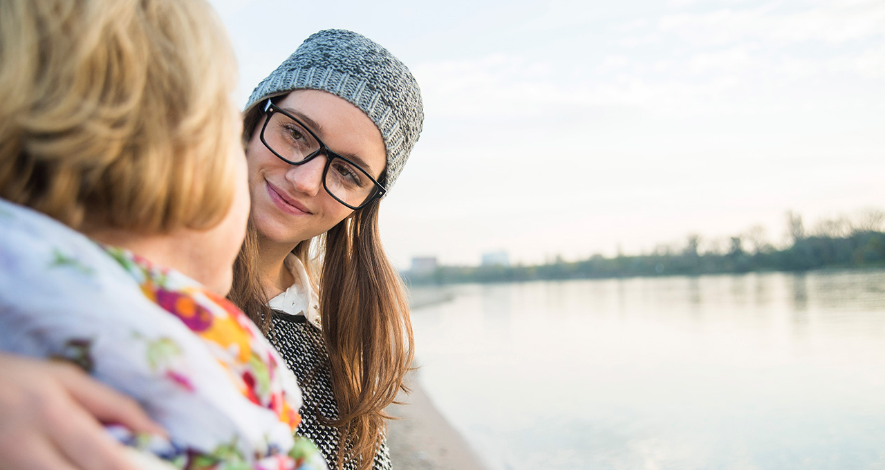 Two women share a moment by a river, one wearing a knitted hat, the other in a floral scarf.