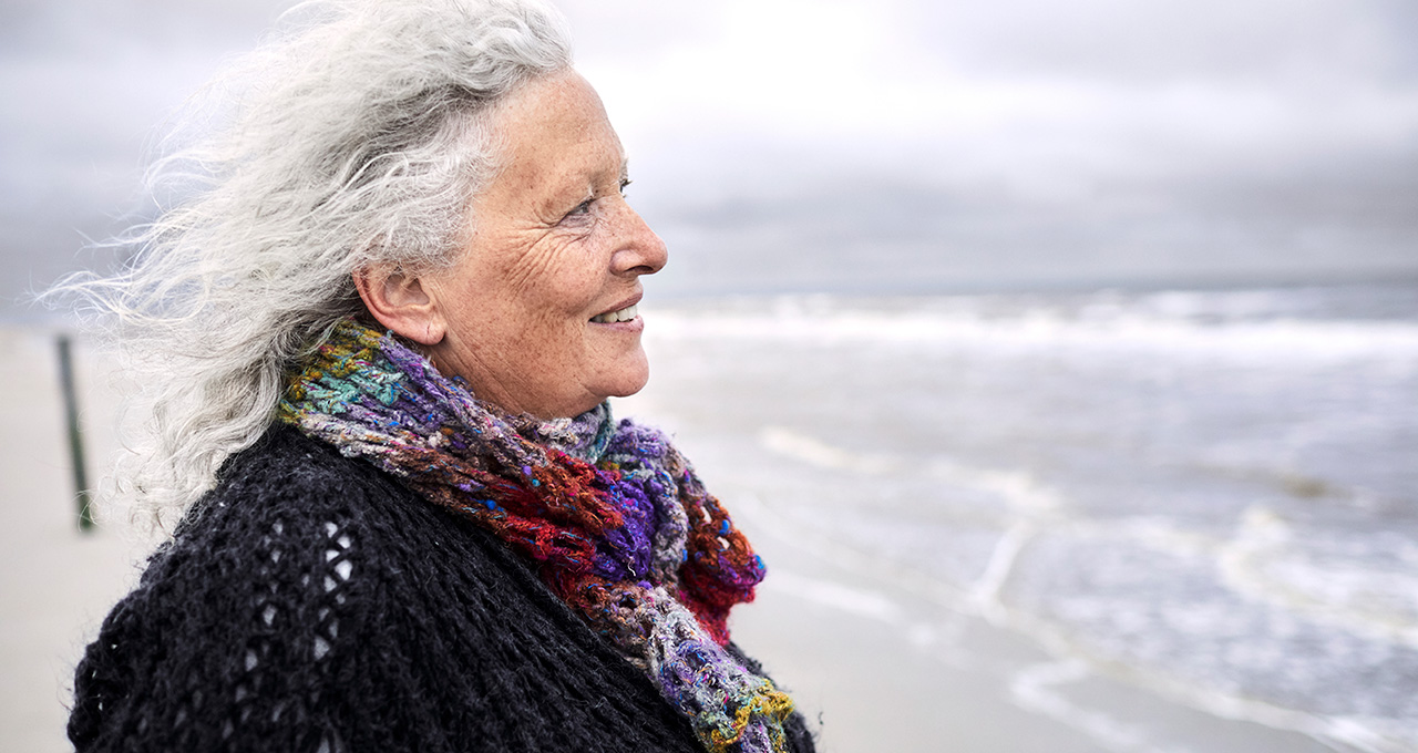 An elderly woman stands by the beach, wearing a colorful scarf against the wind
