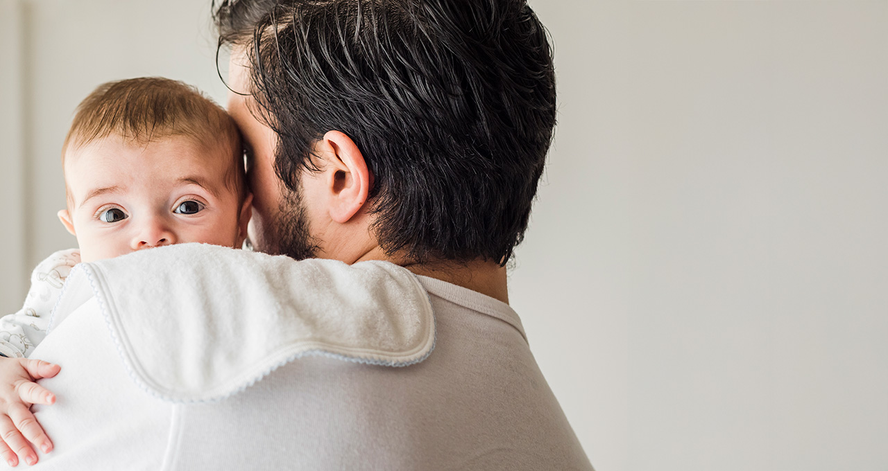 A father holds a child close, both smiling, in a cozy indoor setting with soft lighting.